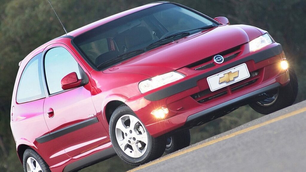 Foto frontal de um Celta 2006 vermelho com os faróis acesos