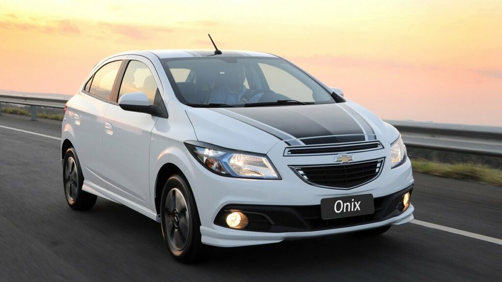 Foto de um Onix 2013 andando em uma pista, o carro possui a cor branca com uma faixa preta no meio