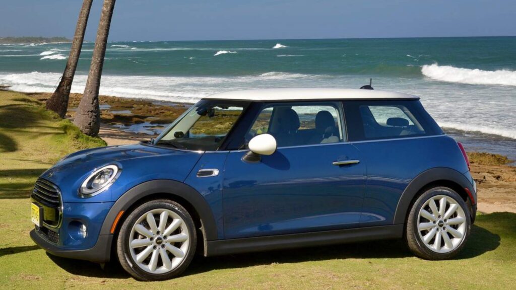MINI COOPER 2015 azul marinho na diagonal em um gramado com a praia no fundo.