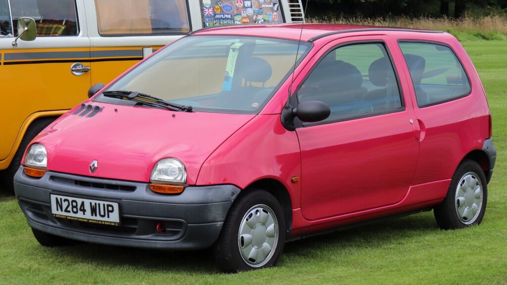Renault Twingo 1995 vermelho parado na diagonal em cima de um gramado do lado de uma Kombi.