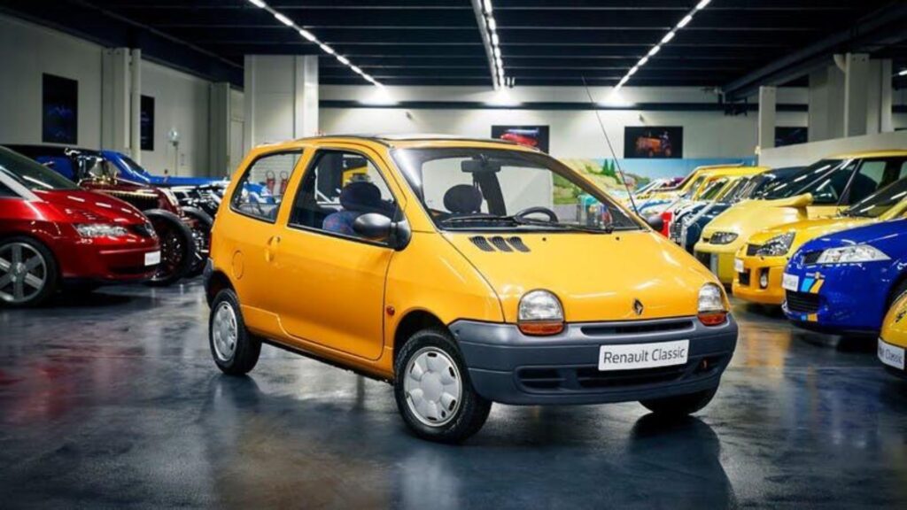 Renault Twingo 1997 amarelo parado na diagonal dentro de uma garagem cheio de carros.