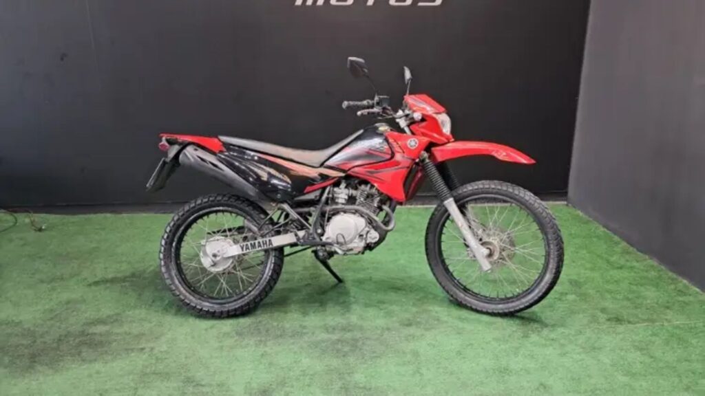 Lateral da YAMAHA XTZ 125 2013 vermelha parada com um fundo preto e em cima de um gramado.