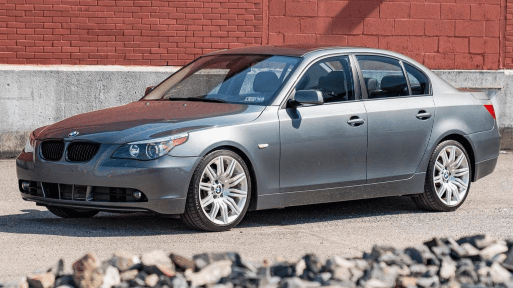 BMW 530i/iA 2004 cinza em parado na diagonal com um galpão de tijolo no fundo.